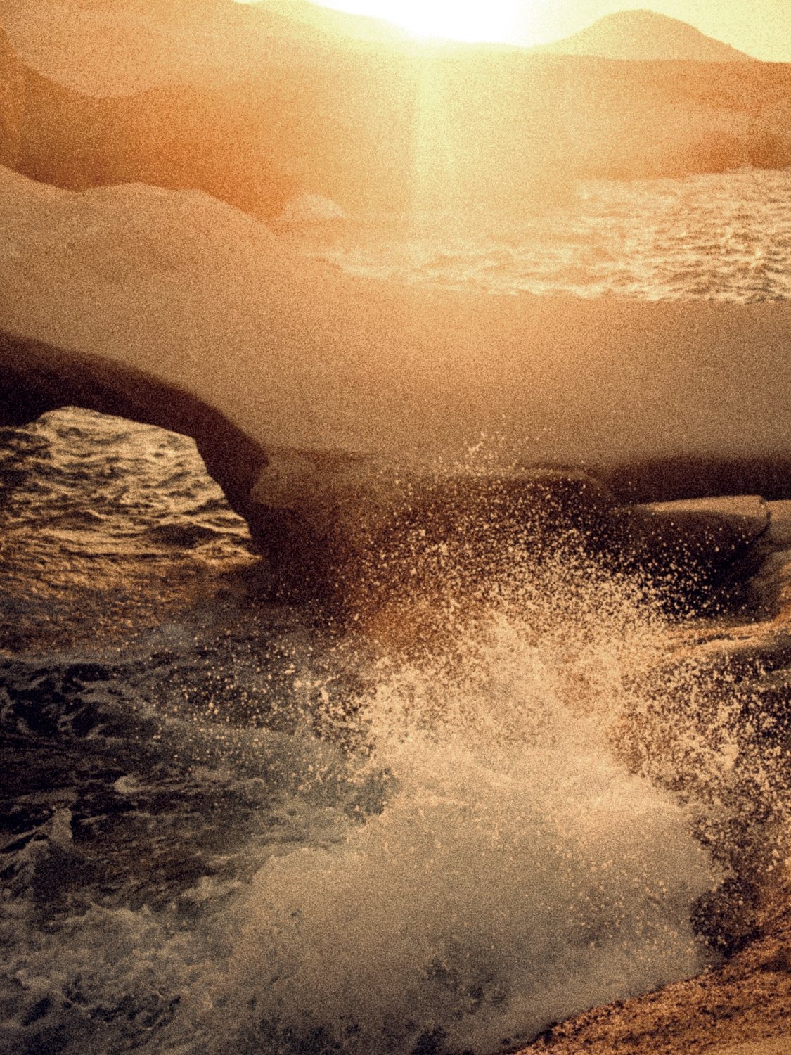 A wave breaking under a white rock arch in the late sun