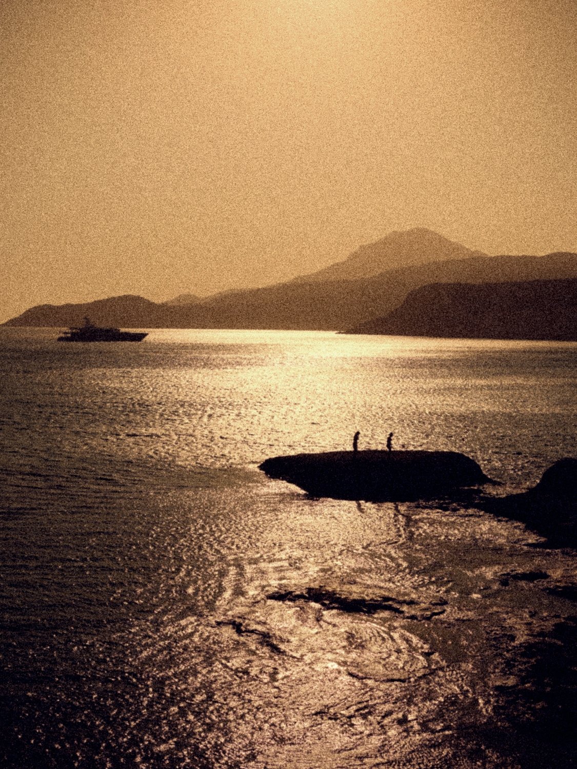 Two figures on a rock in the sea at sunset, a yacht and the island's mountains beyond