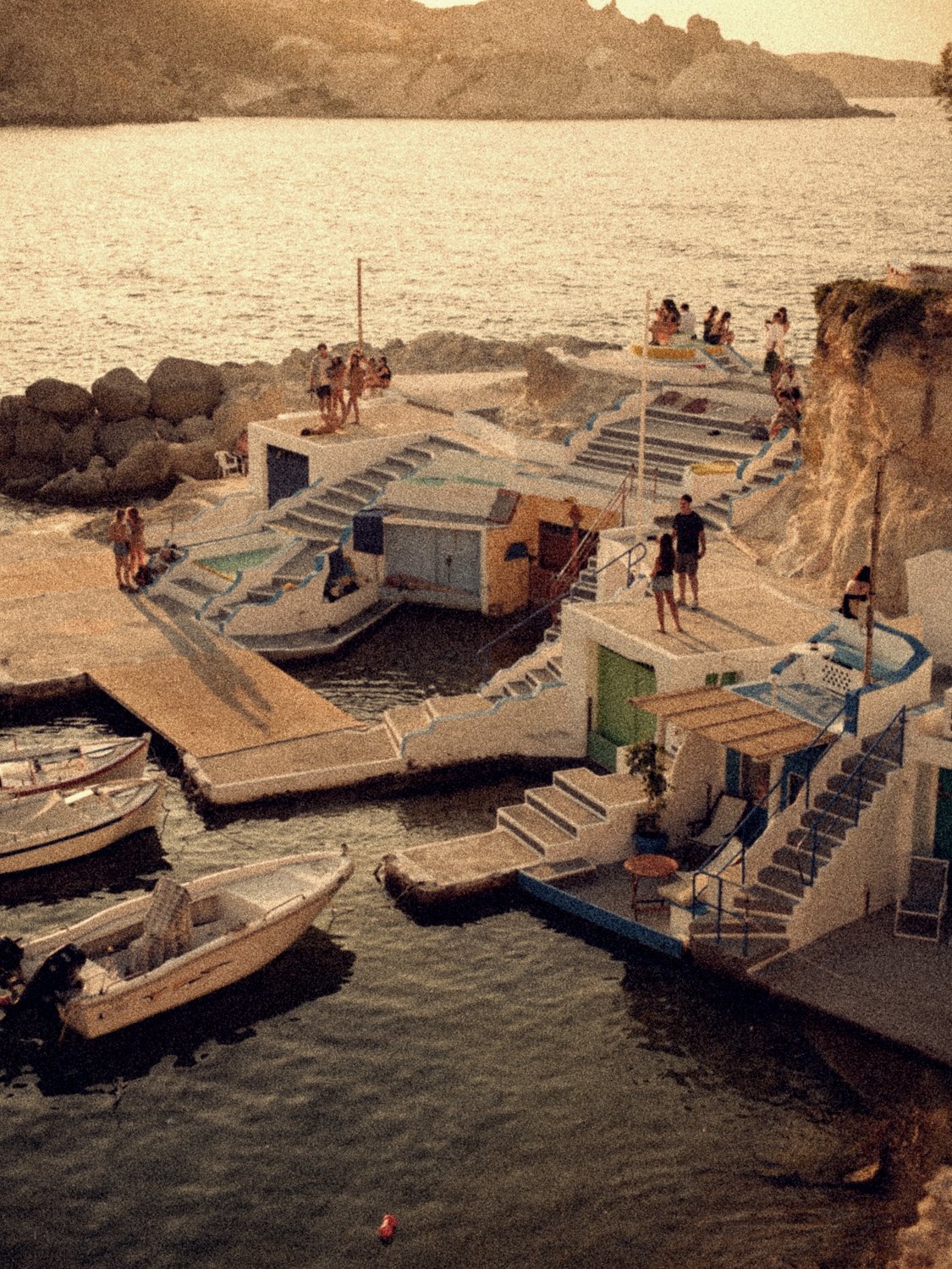 The syrmata of Mandrakia at golden hour: whitewashed boathouses and stairs at the water's edge, people on the rooftops