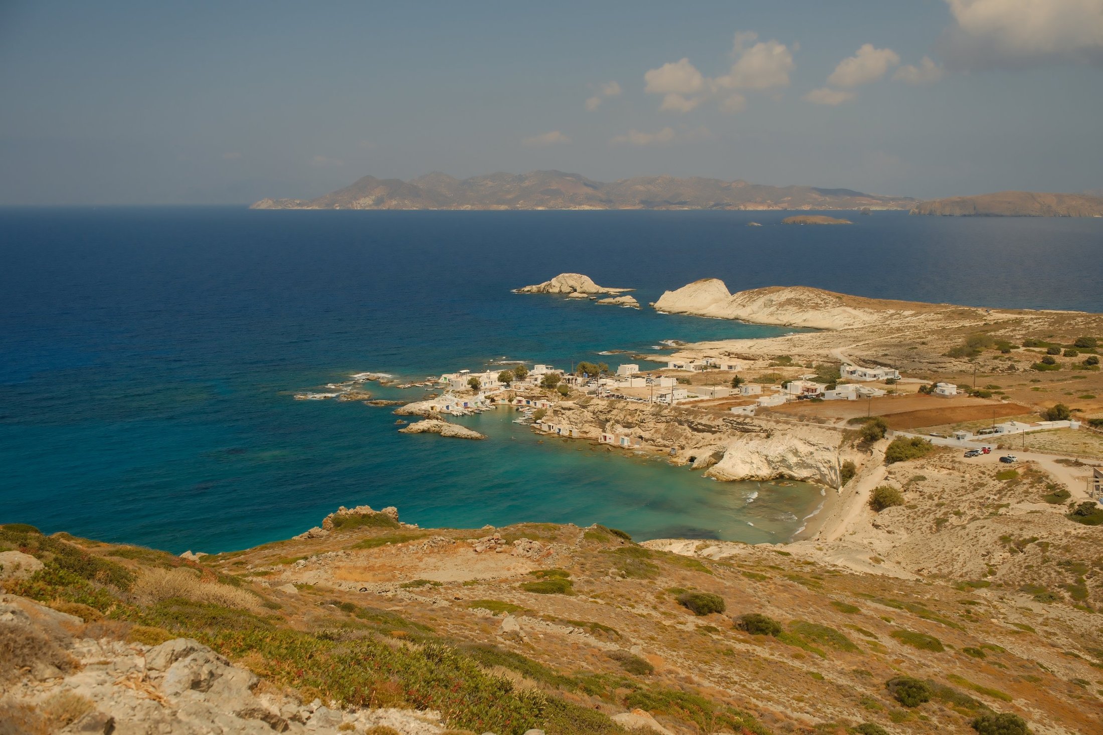 Photograph from the plot: the hillside dropping to Mandrakia bay, the village and its boathouses, white rocks and the open sea, islands on the horizon